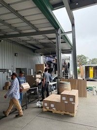 Workers loading boxes onto a truck under a covered loading dock. Outdoors, cloudy day.