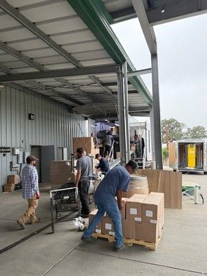 Workers loading boxes onto a truck under a covered loading dock. Gray building, outdoors.