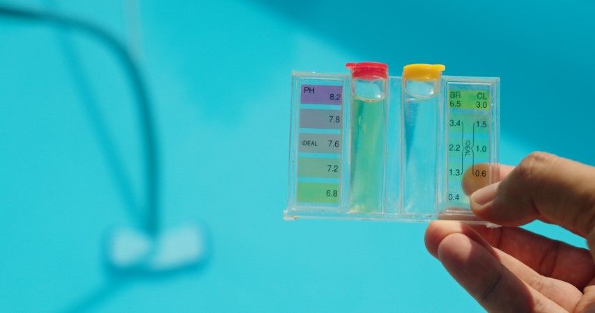 A hand holds a swimming pool water test kit in front of a blue pool; the pH side shows a light green color.