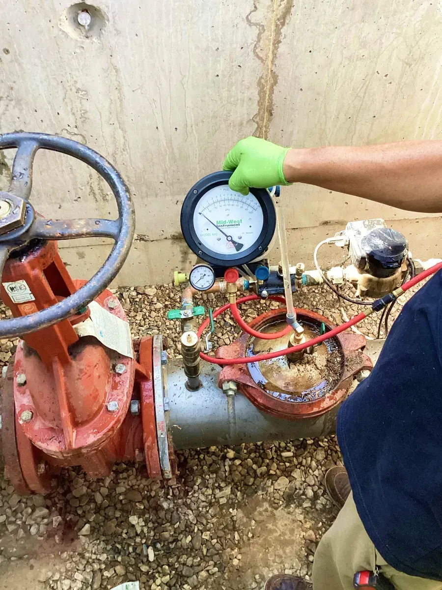 Technician holding a pressure gauge beside exposed plumbing valves and pipes against a wall