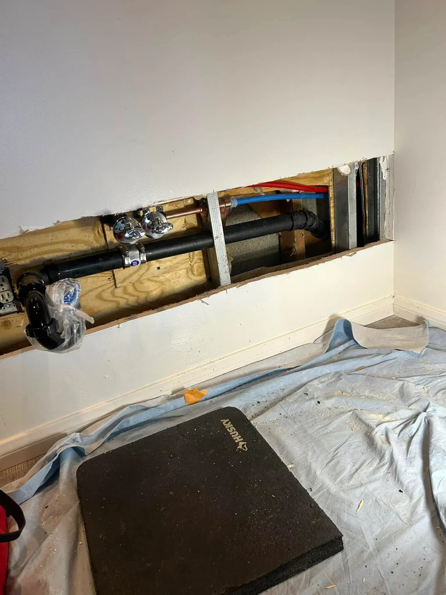 Damaged wall with exposed wiring above a bed covered in a gray sheet, with a black cushion in the foreground