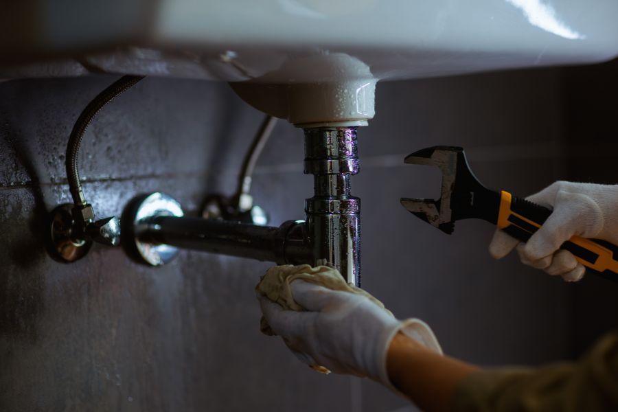 Gloved hands use a wrench to repair a sink drain under a bathroom sink.