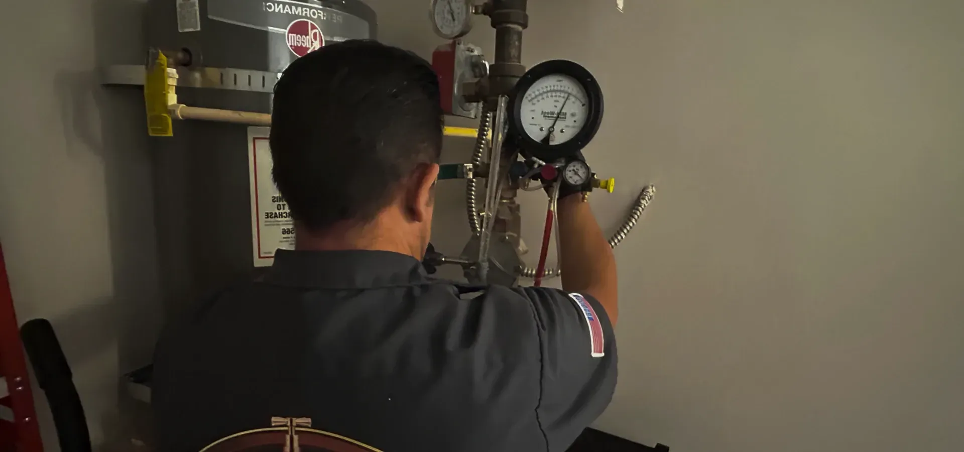 Person inspecting a wall-mounted pressure gauge and pipes in a utility room.
