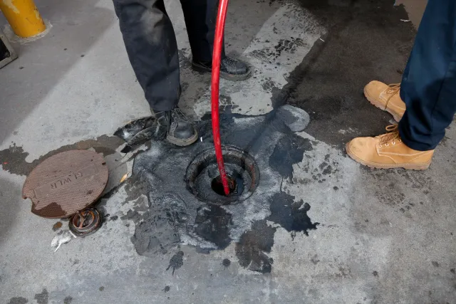 Workers using a red hose to clean a dark spill around a floor drain on concrete.