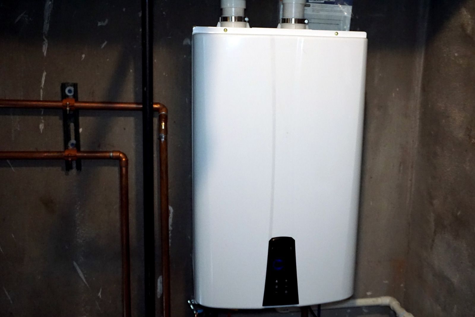 White wall-mounted water heater in a basement, with copper pipes and dark concrete walls