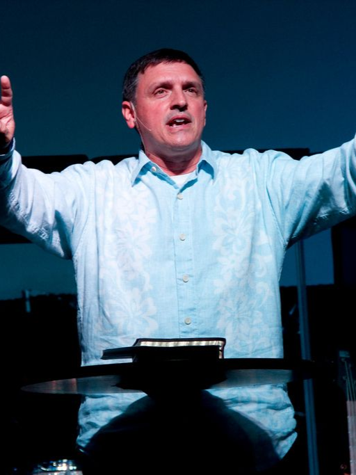 Man speaking with arms raised, wearing a light blue shirt. Standing behind a dark podium, indoors.