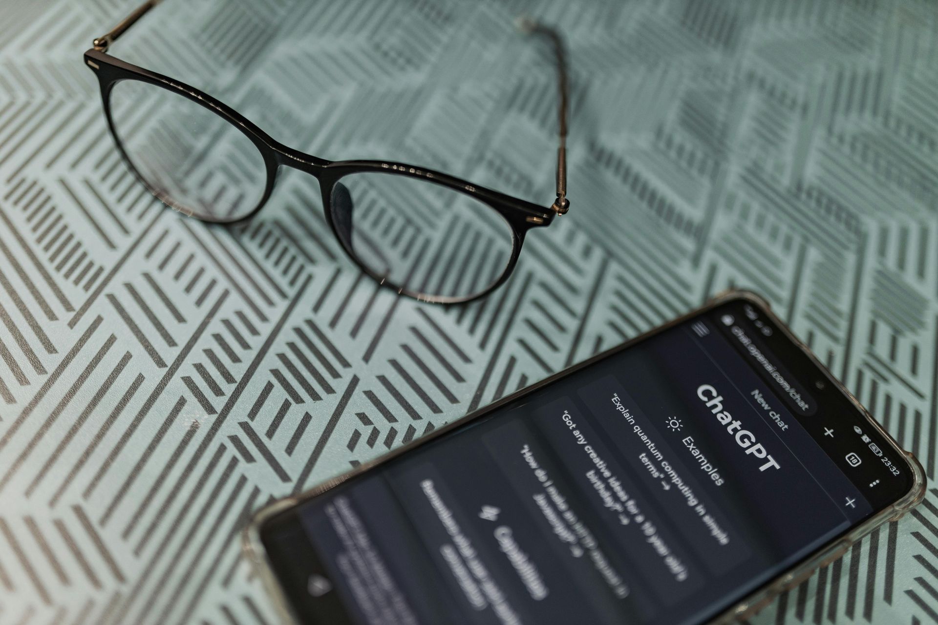 Black-framed glasses and smartphone on a patterned surface; smartphone displays the ChatGPT interface.