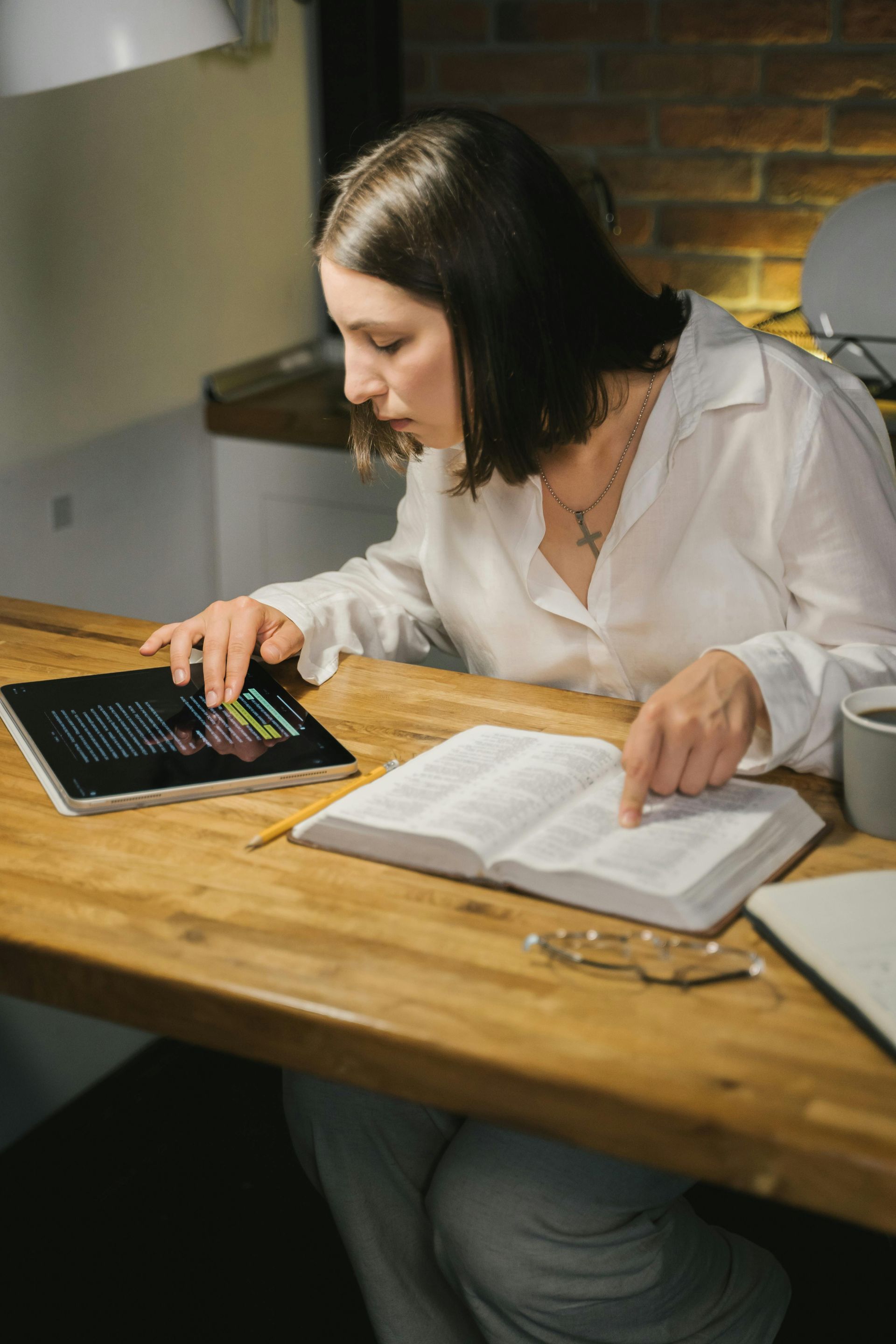 A woman is sitting at a table reading a book and using a tablet.