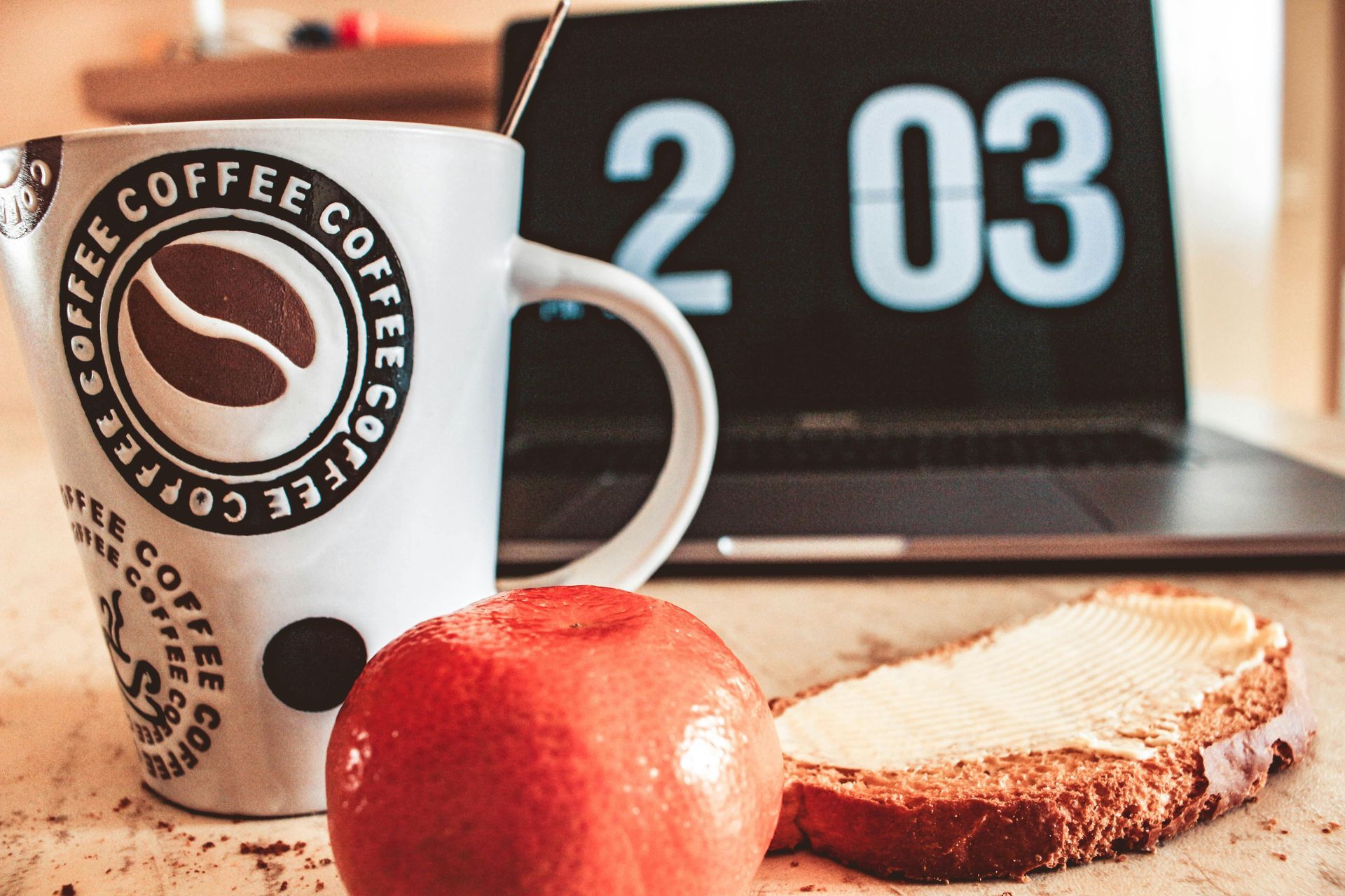 White coffee mug with coffee bean logo, red apple, toast, and laptop displaying time 2:03 on a wooden table.
