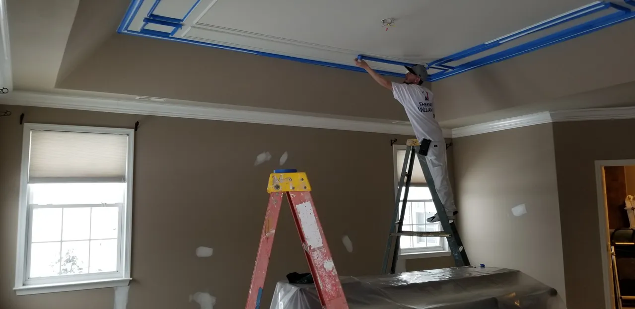Person on a ladder tapes the ceiling trim for painting. Room has a window, walls are brown.