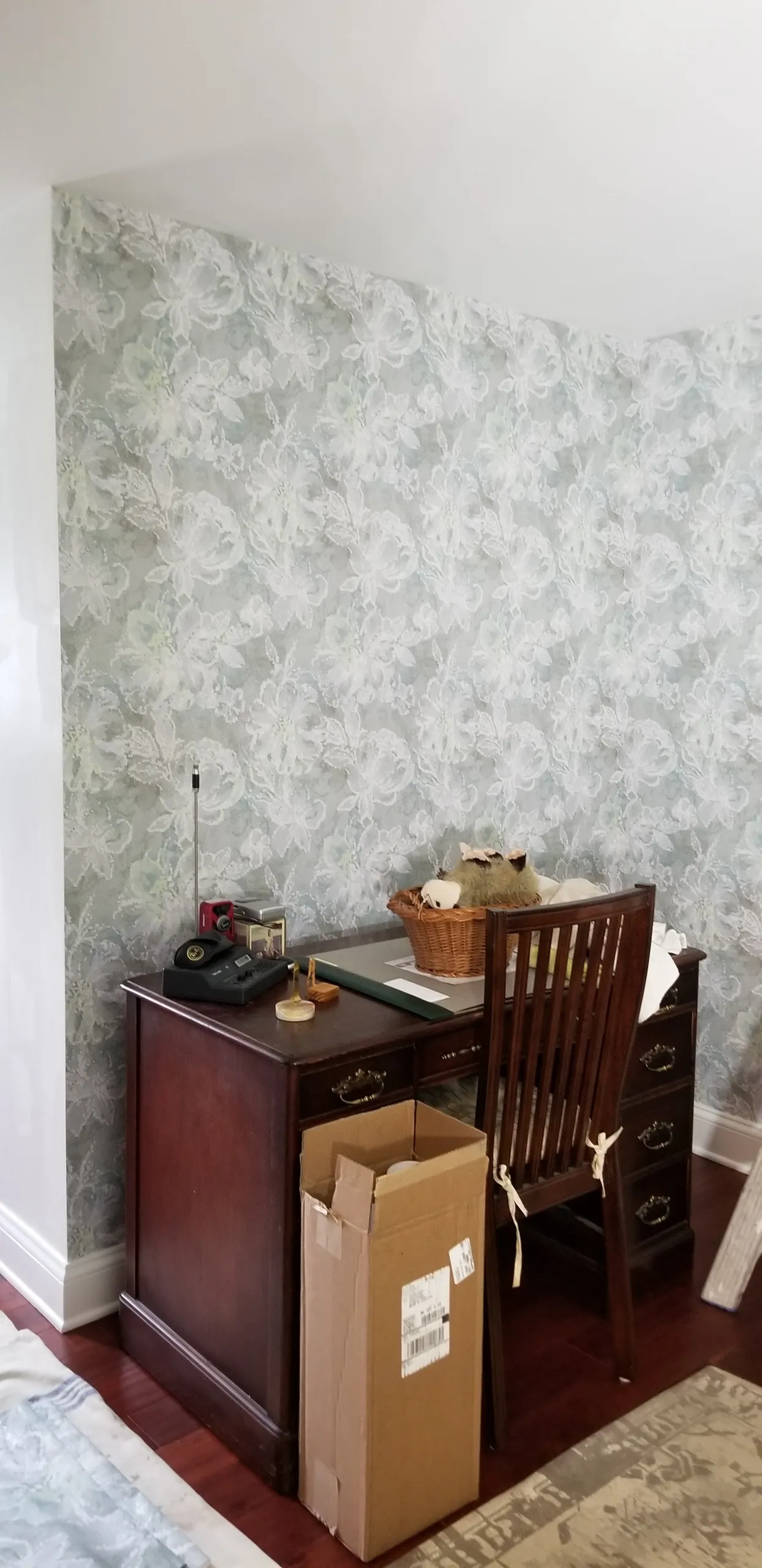 A room with patterned wallpaper, a dark wooden desk, and a chair. A cardboard box sits in front of the desk.