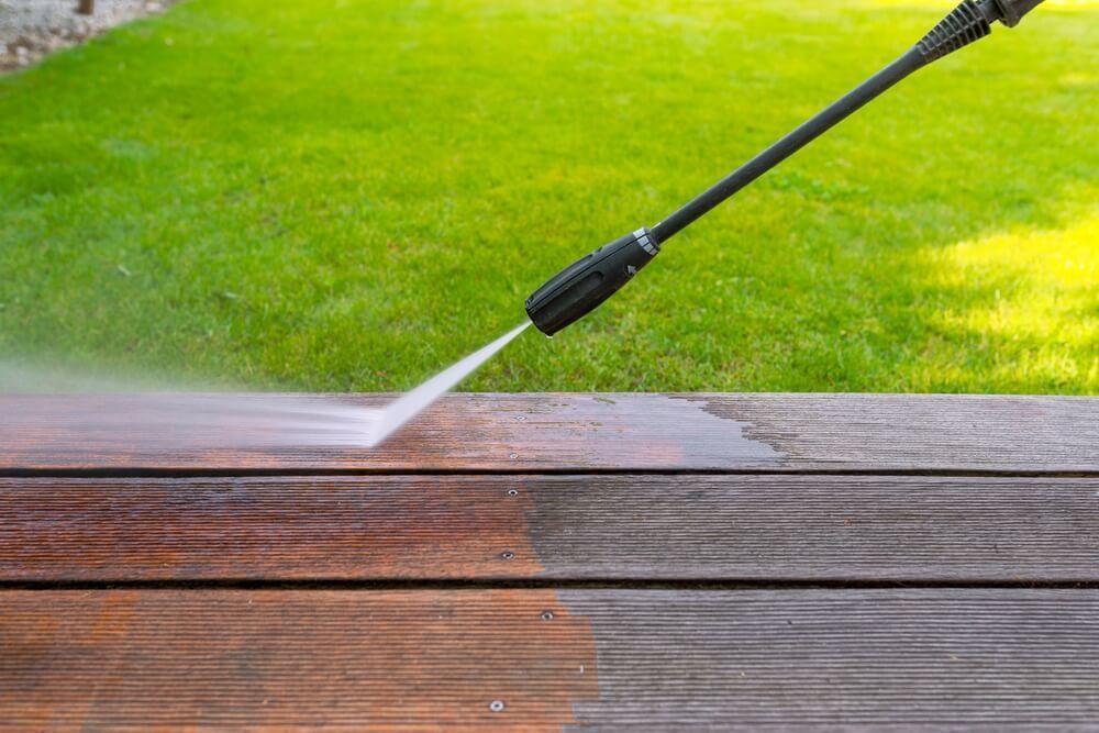 Pressure washer cleaning a wooden deck; showing before and after cleaning effect; green grass in background.
