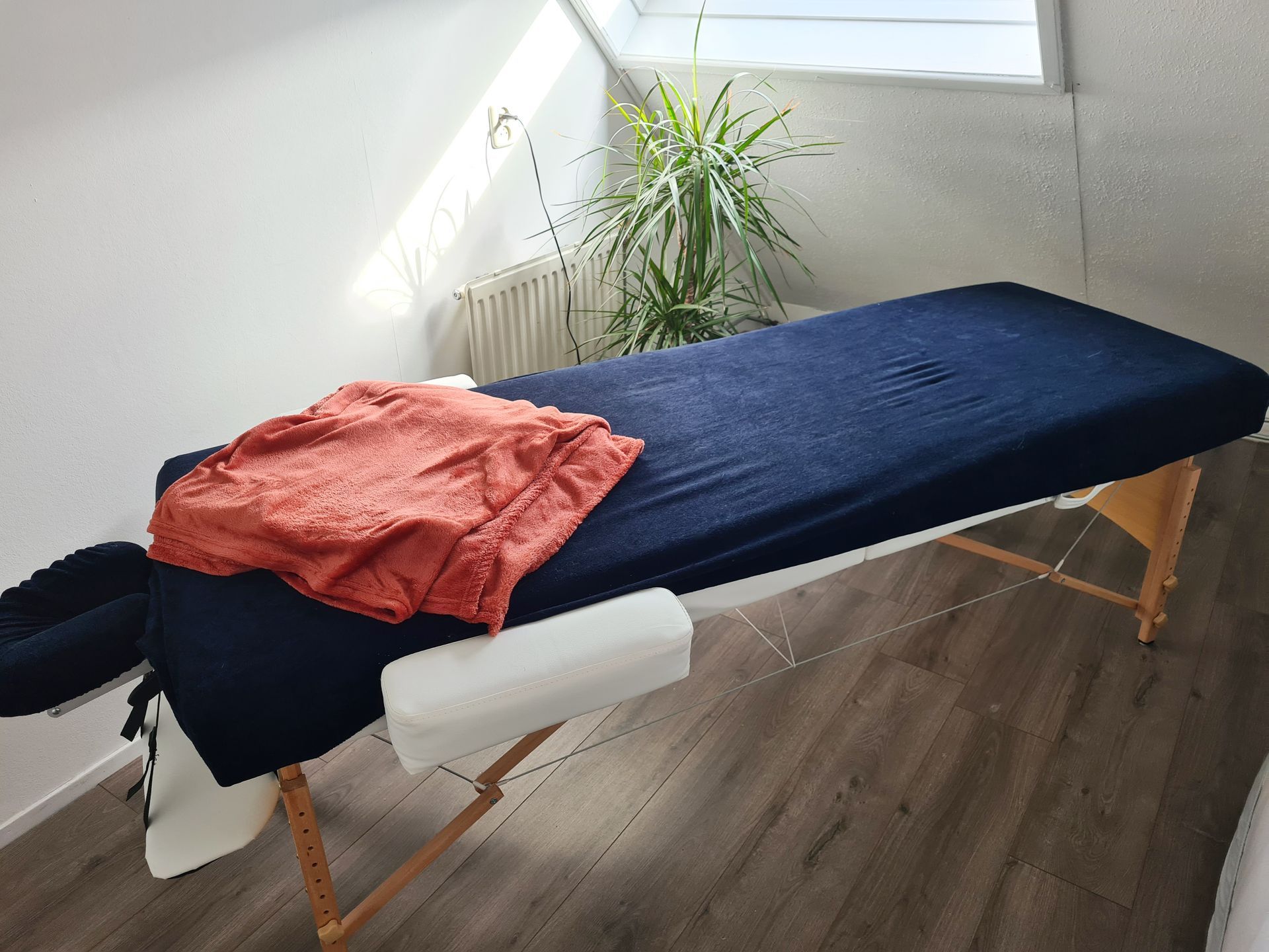Massagetafel met blauwe hoes, oranje handdoek en hoofdsteun, in een kamer met een plant en een raam.