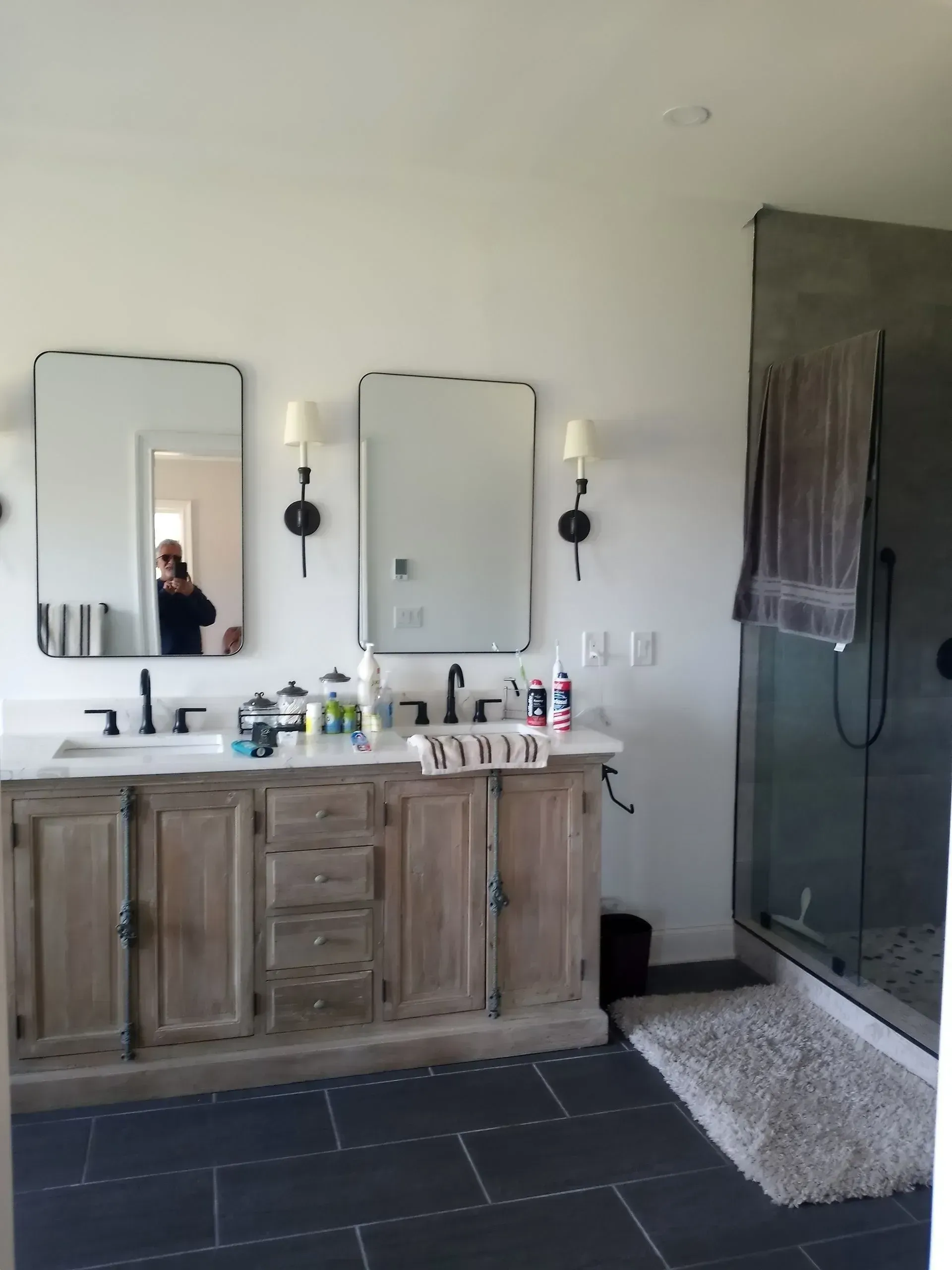 Bathroom with double vanity, mirrors, sconces, and a glass shower with a gray rug