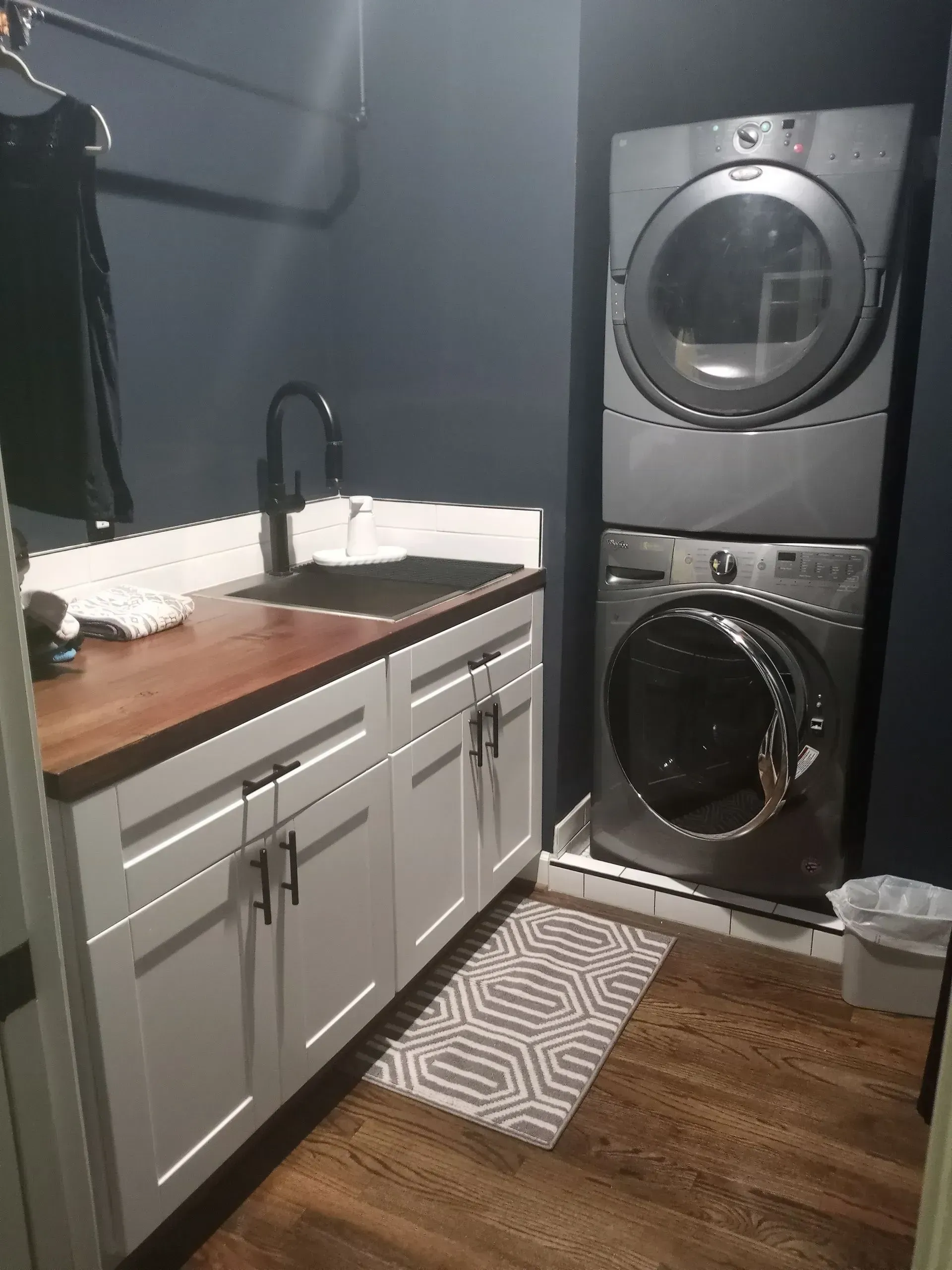 Small laundry room with stacked washer and dryer beside a white sink cabinet and wood countertop.