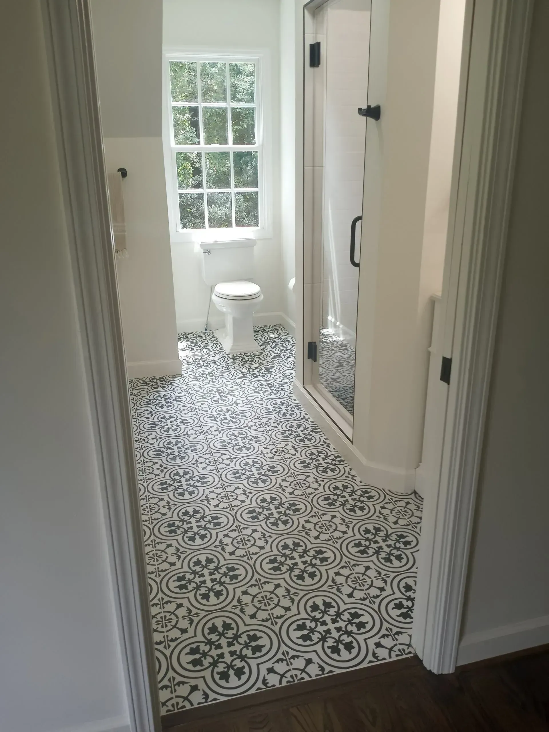 Narrow bathroom with black-and-white patterned tile floor, white walls, shower, toilet, and window.