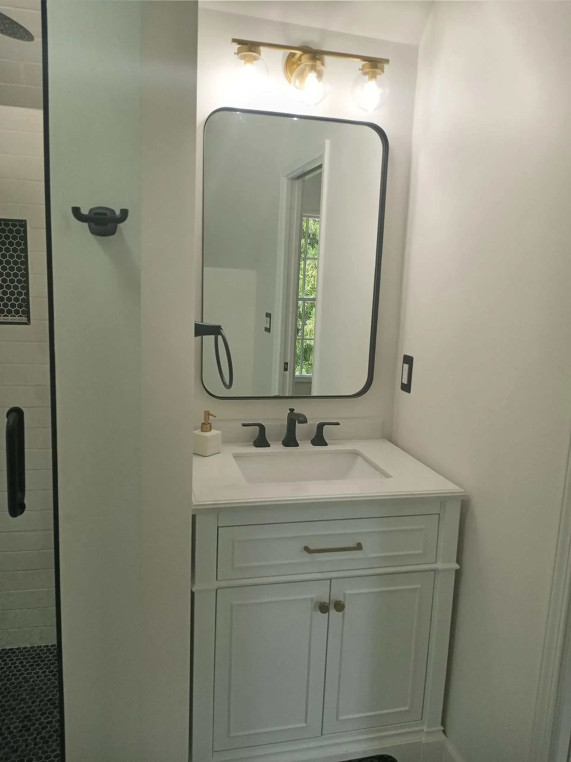 White bathroom vanity with mirror, wall sconces, sink, and a window reflecting greenery.