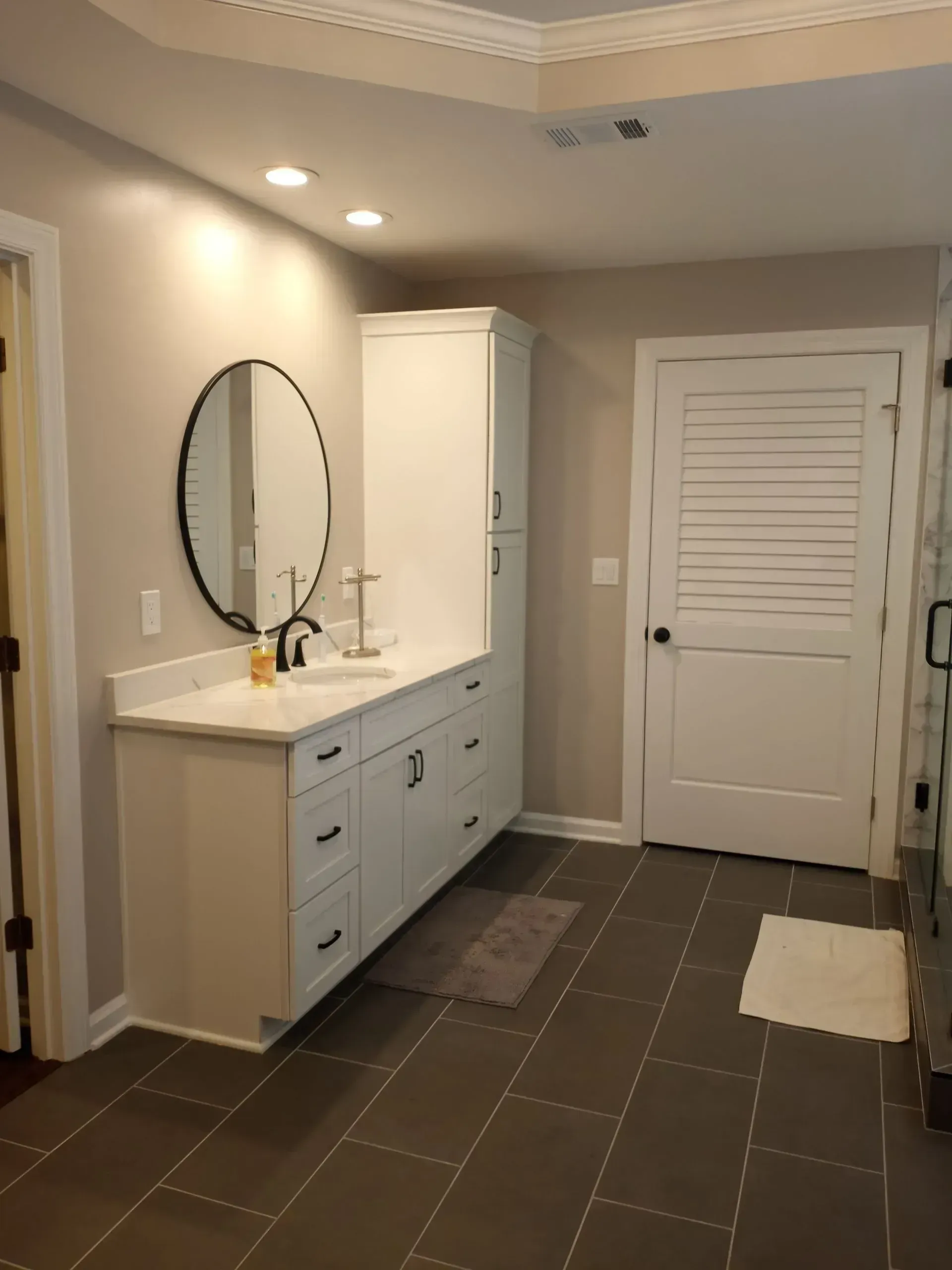 Bathroom vanity with round mirror, white cabinets, and closed door on beige tile floor.