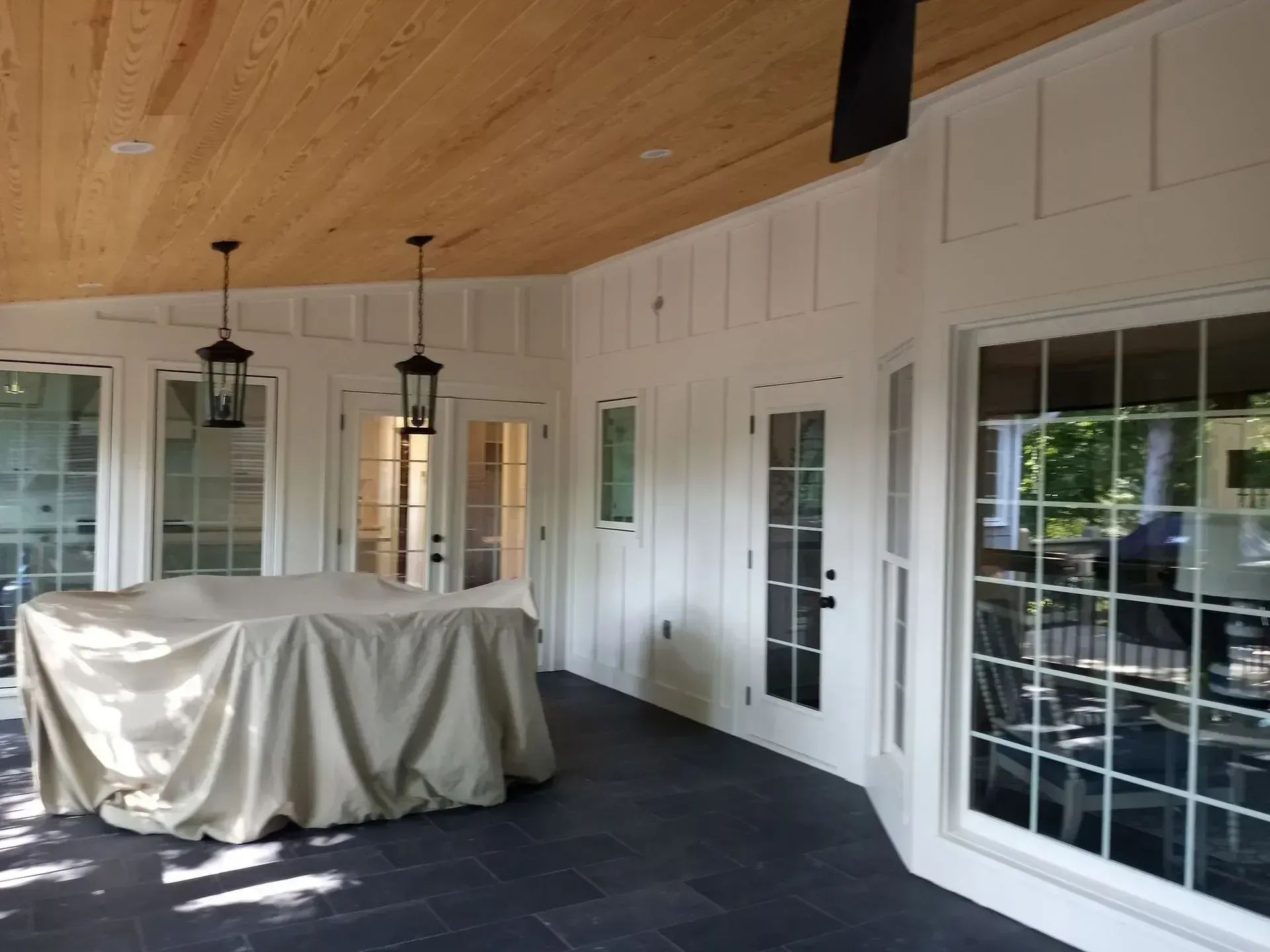 Covered patio with white paneled walls, wood ceiling, hanging lanterns, and a covered grill or table.
