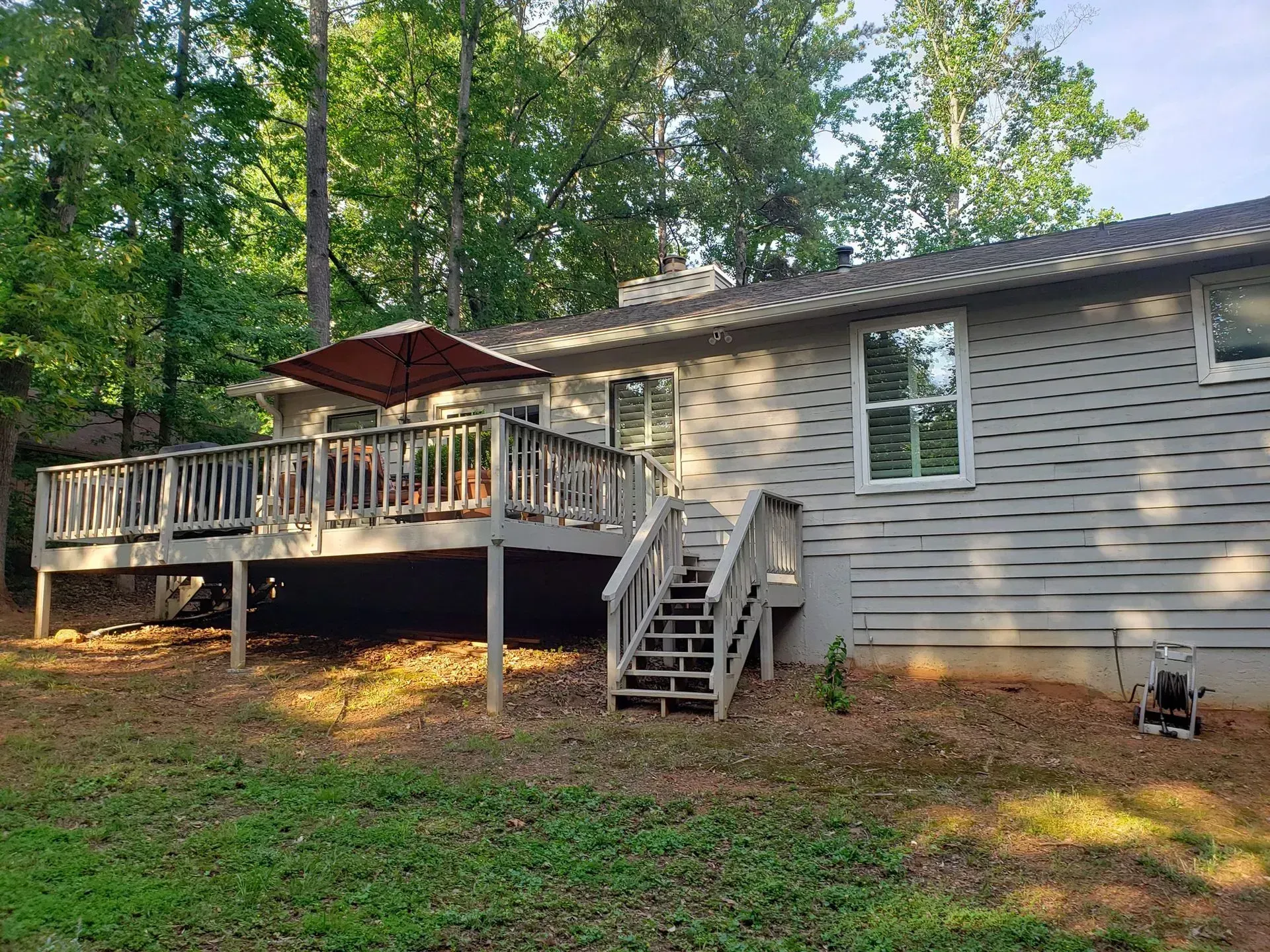 Wooded backyard with a raised deck, red umbrella, and stairs beside a beige house.