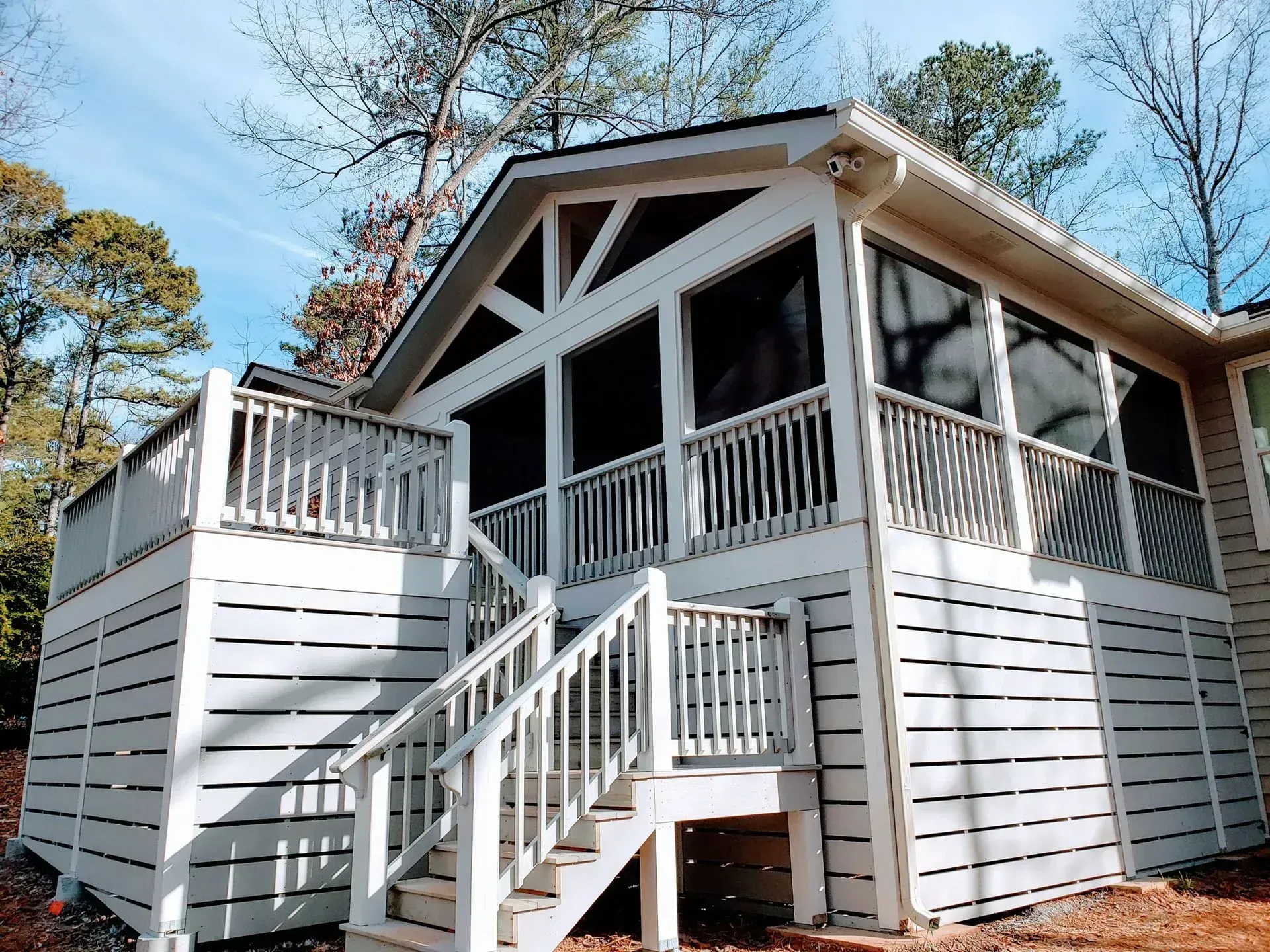 Wooden house deck and stairs with white railings, surrounded by trees under a blue sky