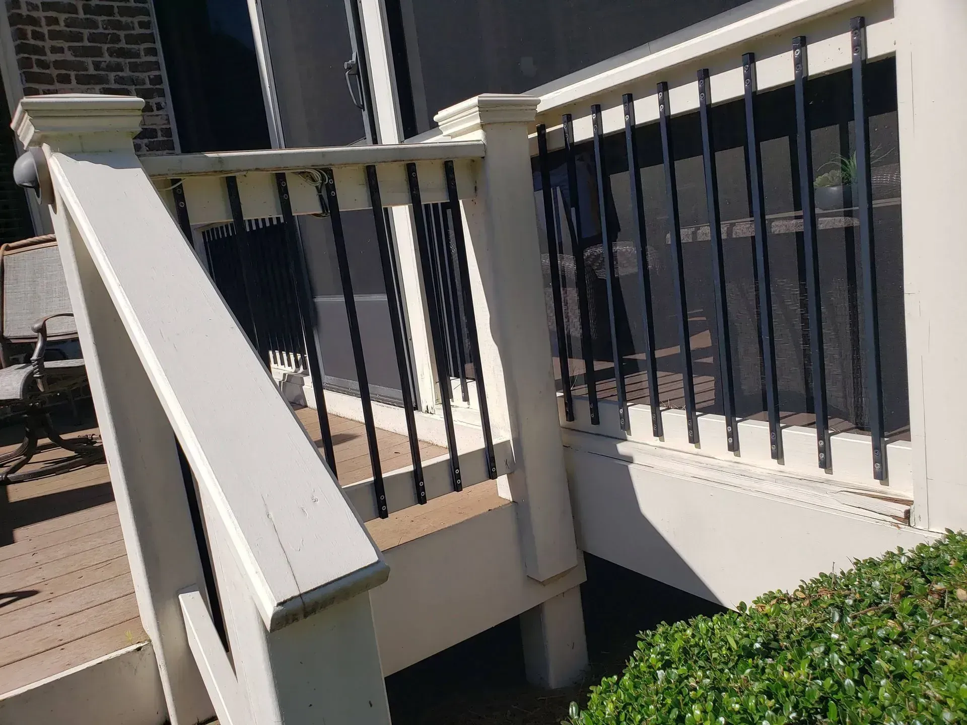 White staircase and railing leading to a deck beside a dark-sided house, with green shrubs below