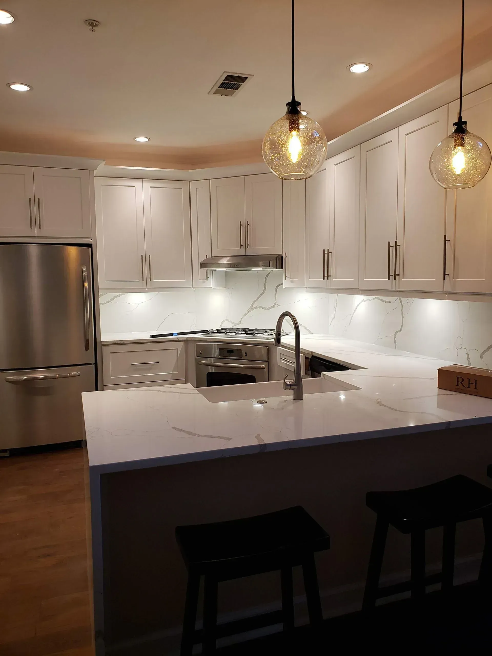 Modern white kitchen with marble island, stainless fridge, pendant lights, and dark stools