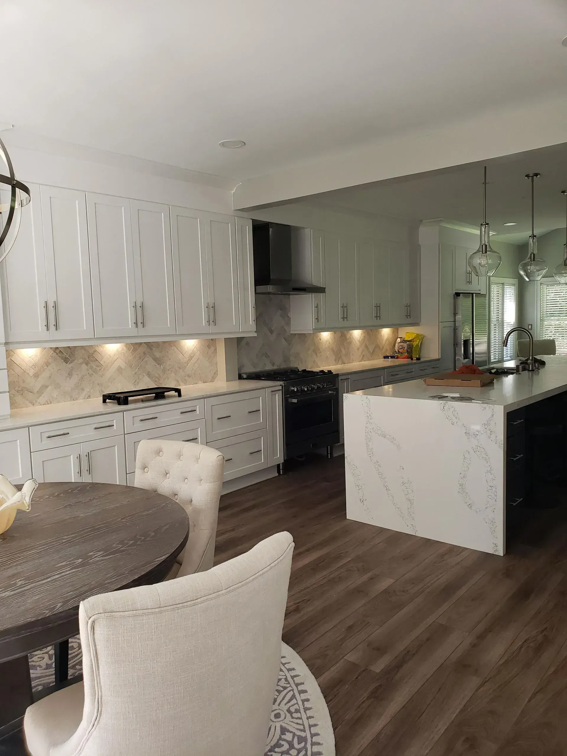Modern white kitchen and dining area with marble island, dark wood floor, and round table with upholstered chairs