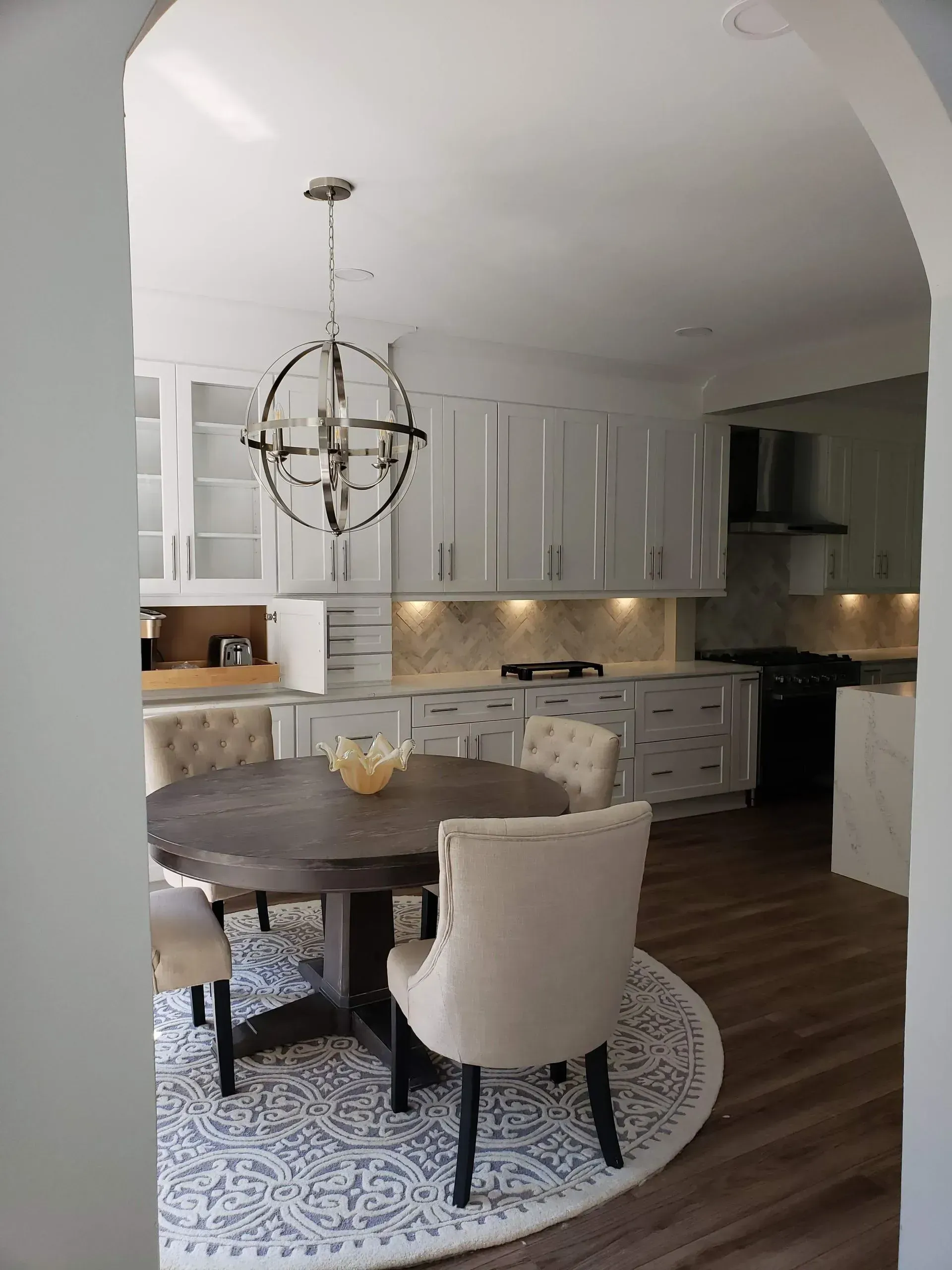 Modern white kitchen and dining area with round table, upholstered chairs, patterned rug, and pendant light