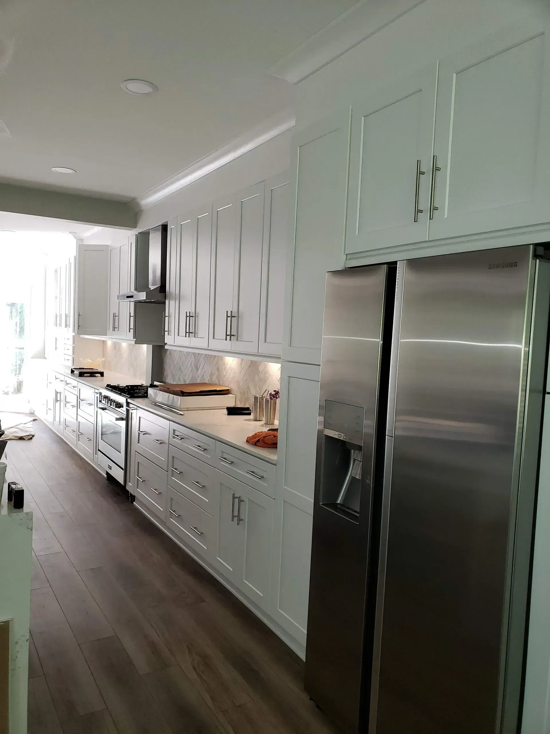 Bright white galley kitchen with stainless steel refrigerator and long counter along a hardwood floor.