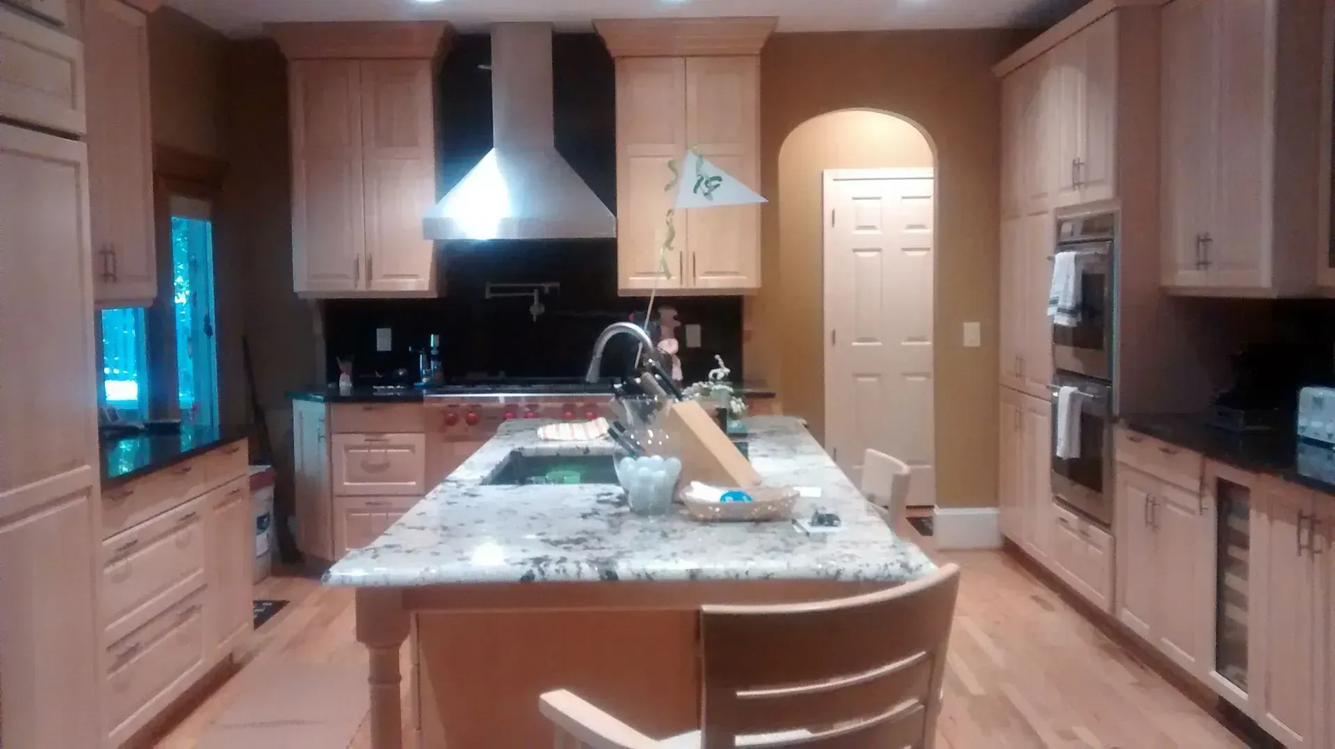 Modern kitchen with light wood cabinets, central island, and white range hood over a dark backsplash.
