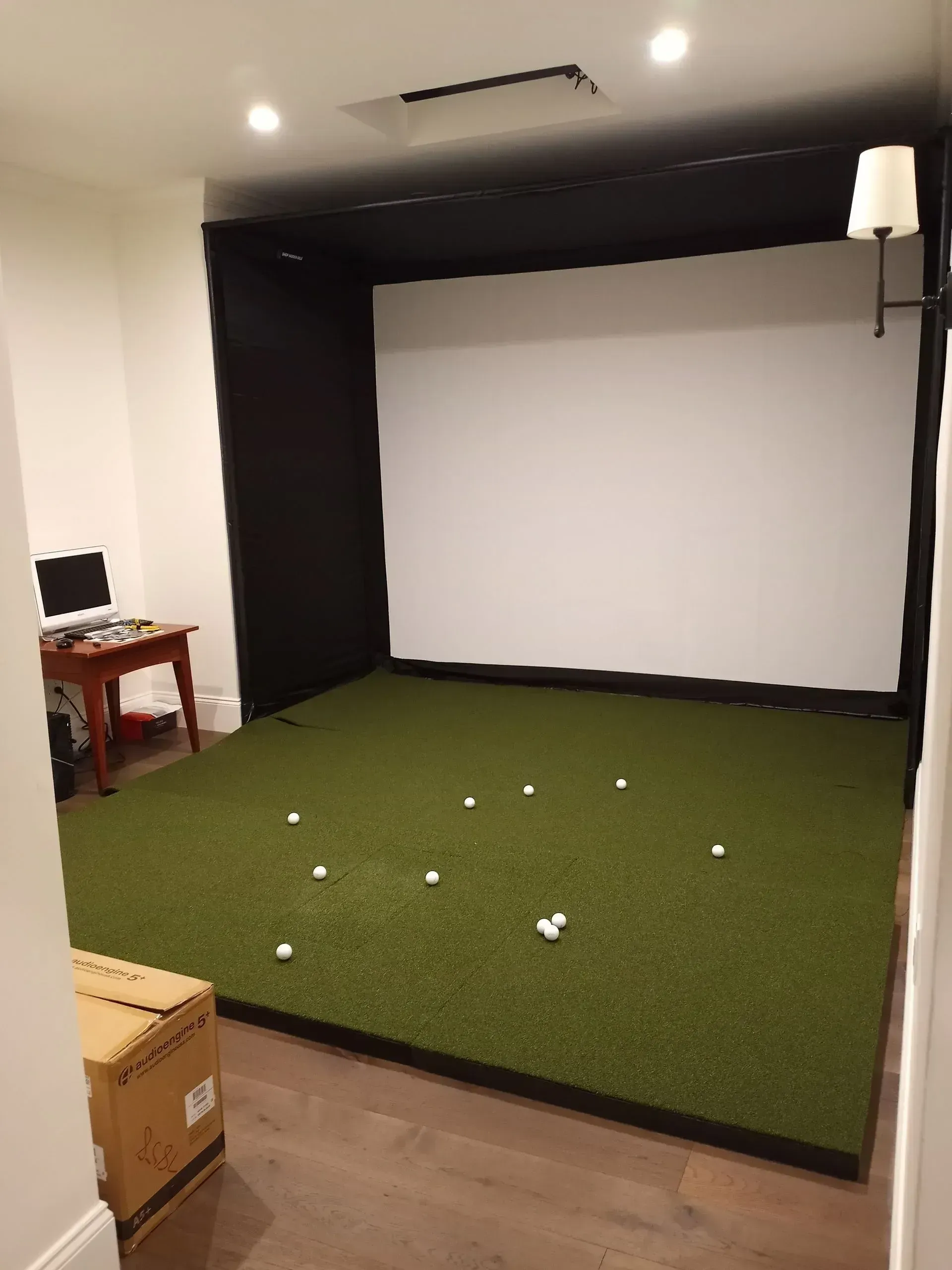 Indoor golf simulator room with green turf, white projection screen, and black enclosure