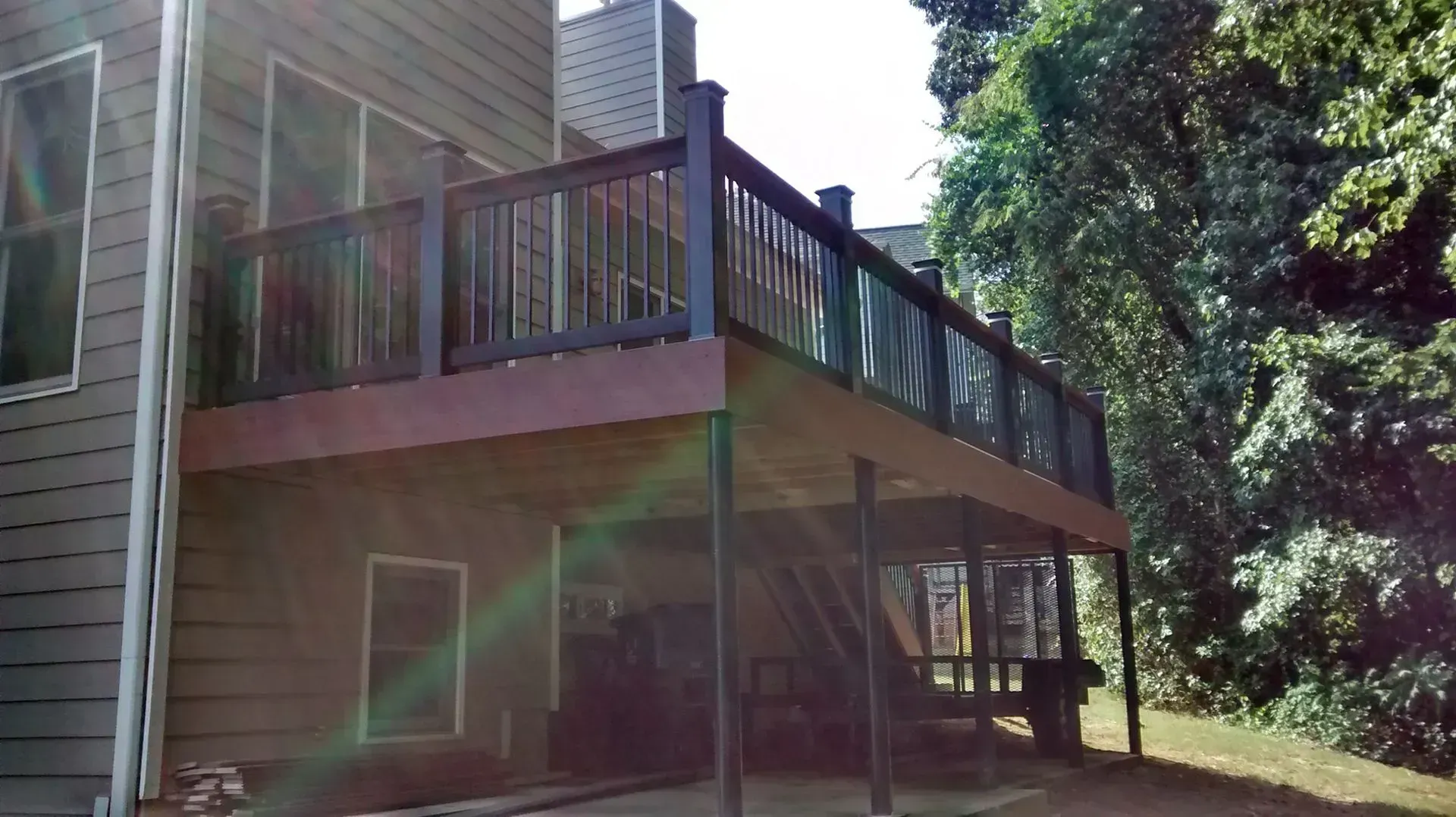 Back deck with dark railing beside house, overlooking a grassy yard and trees in sunlight