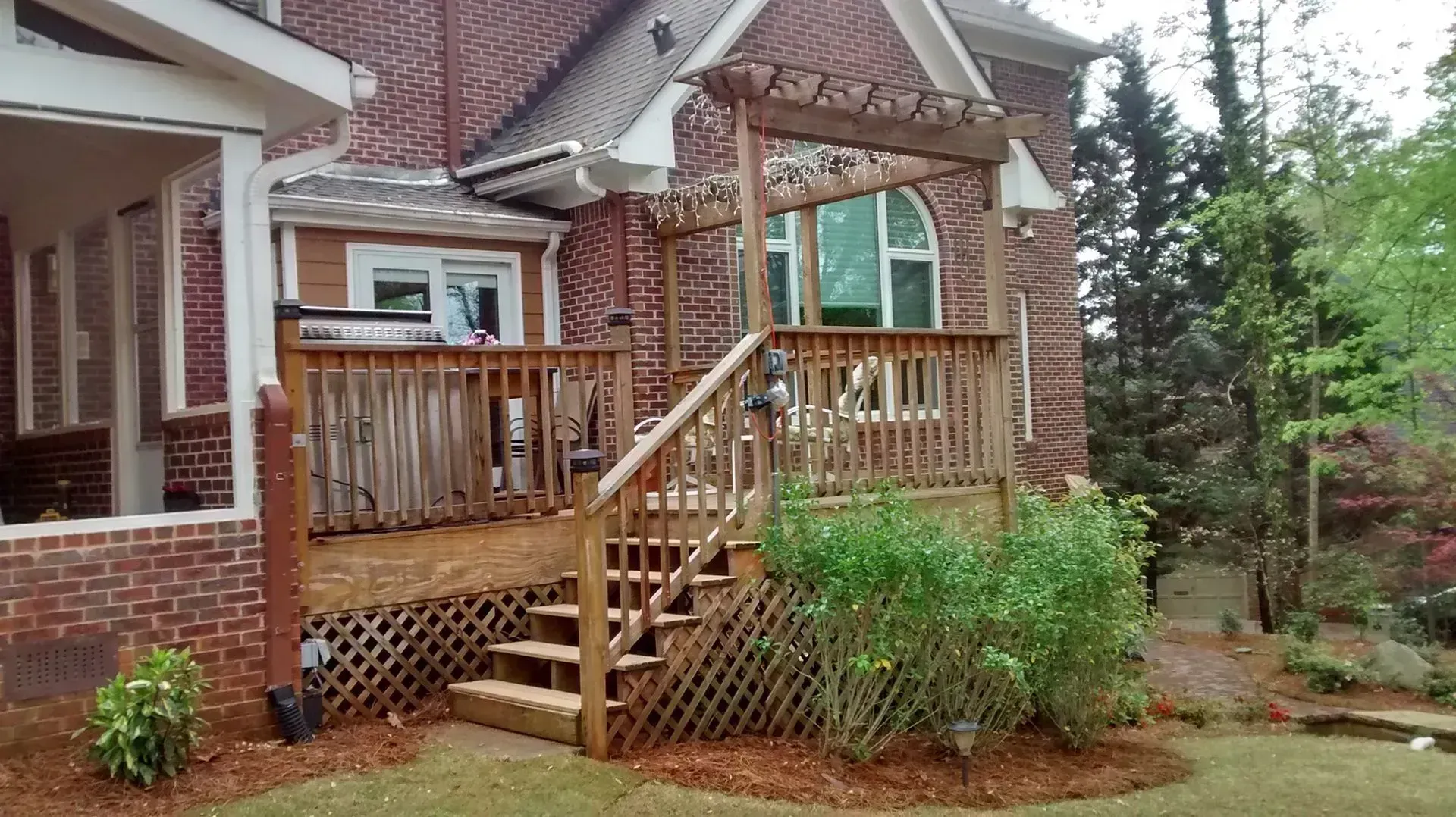 Brick house front porch with wooden railing, stairs, and green shrubs in a wooded yard