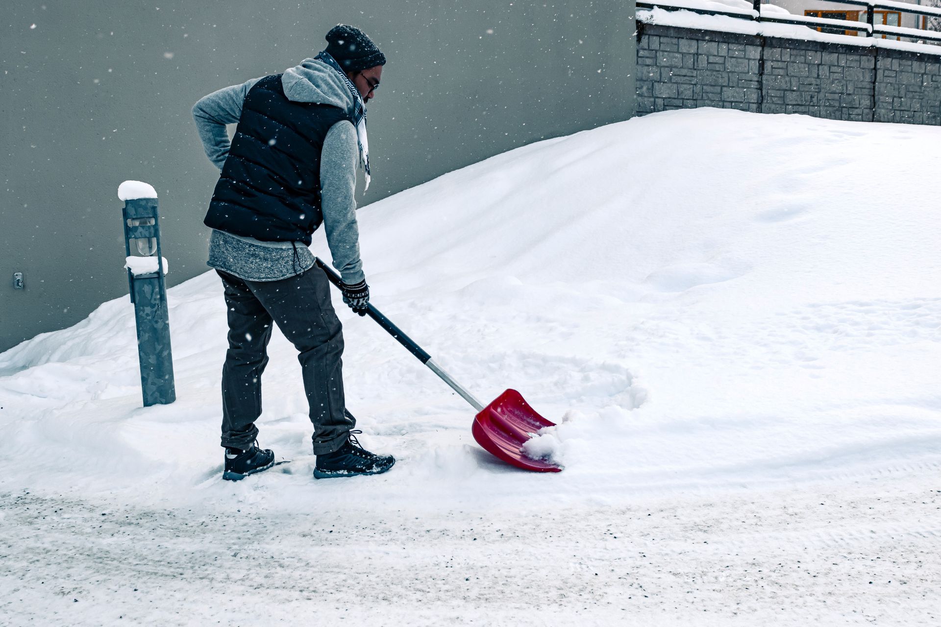 Man shoveling snow from a sidewalk with a red shovel. Gray building in the background, snowy setting.