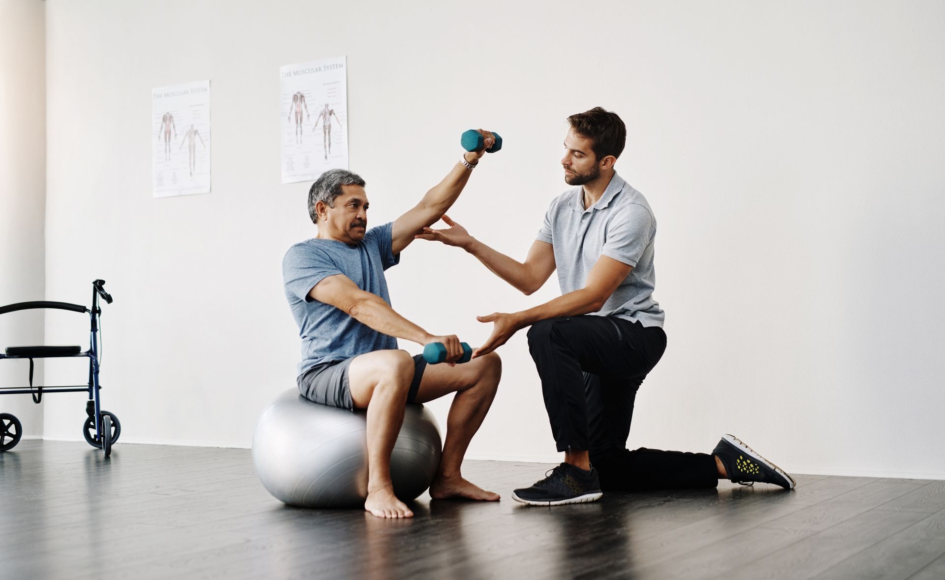 A man is sitting on a pilates ball while a physical therapist helps him lift dumbbells.