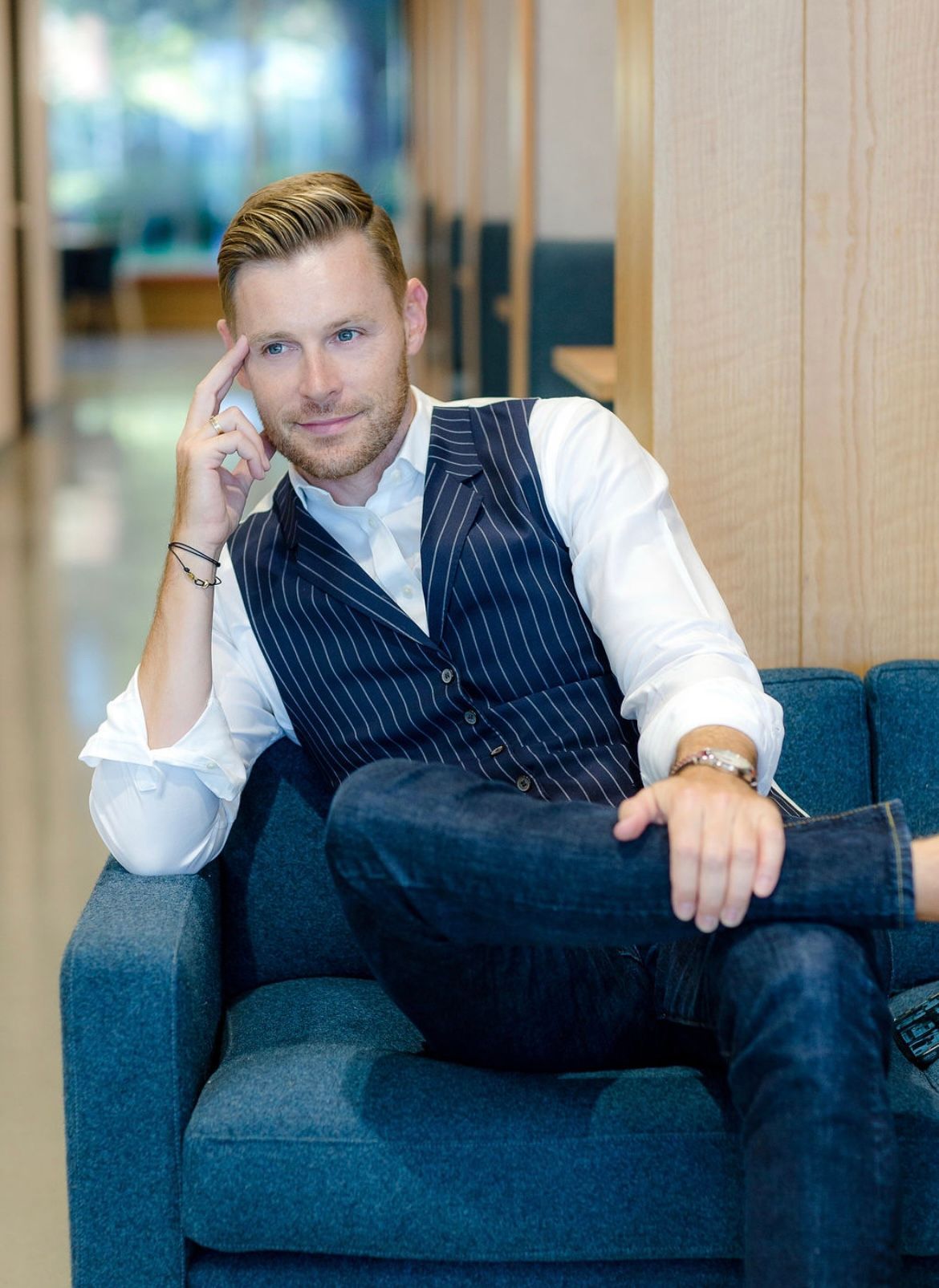 Man in vest and jeans sits on a blue couch, hand on head, thoughtful expression. Modern office setting.