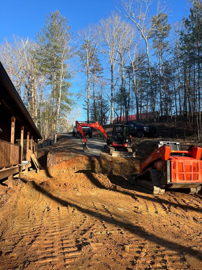 Construction site with excavator and dump truck on a dirt slope beside a building and trees