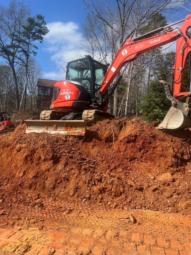 Red excavator working on a dirt mound in a wooded area under a blue sky