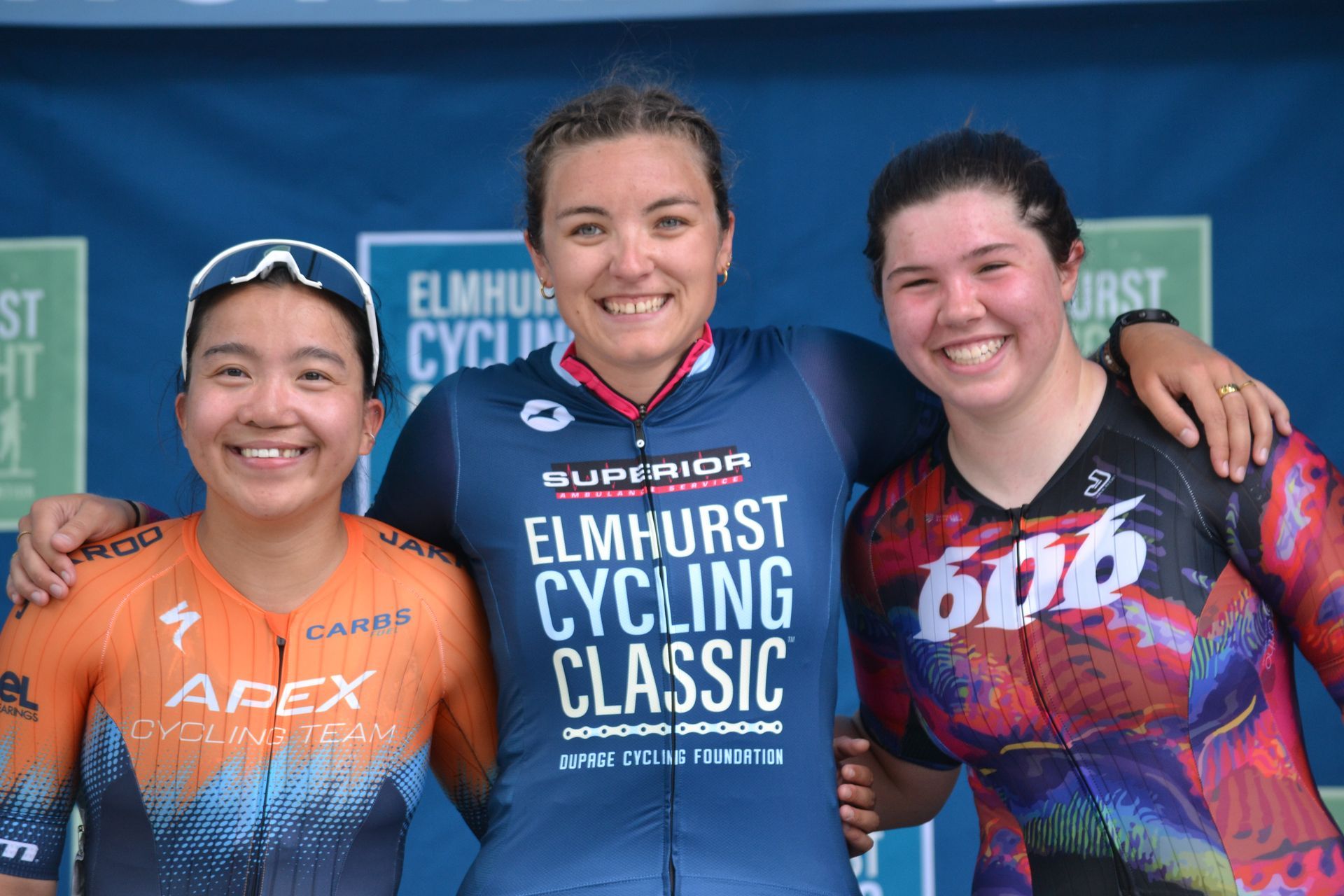 Three women are posing for a picture together and one of them is wearing a jersey that says elmhurst cycling classic.