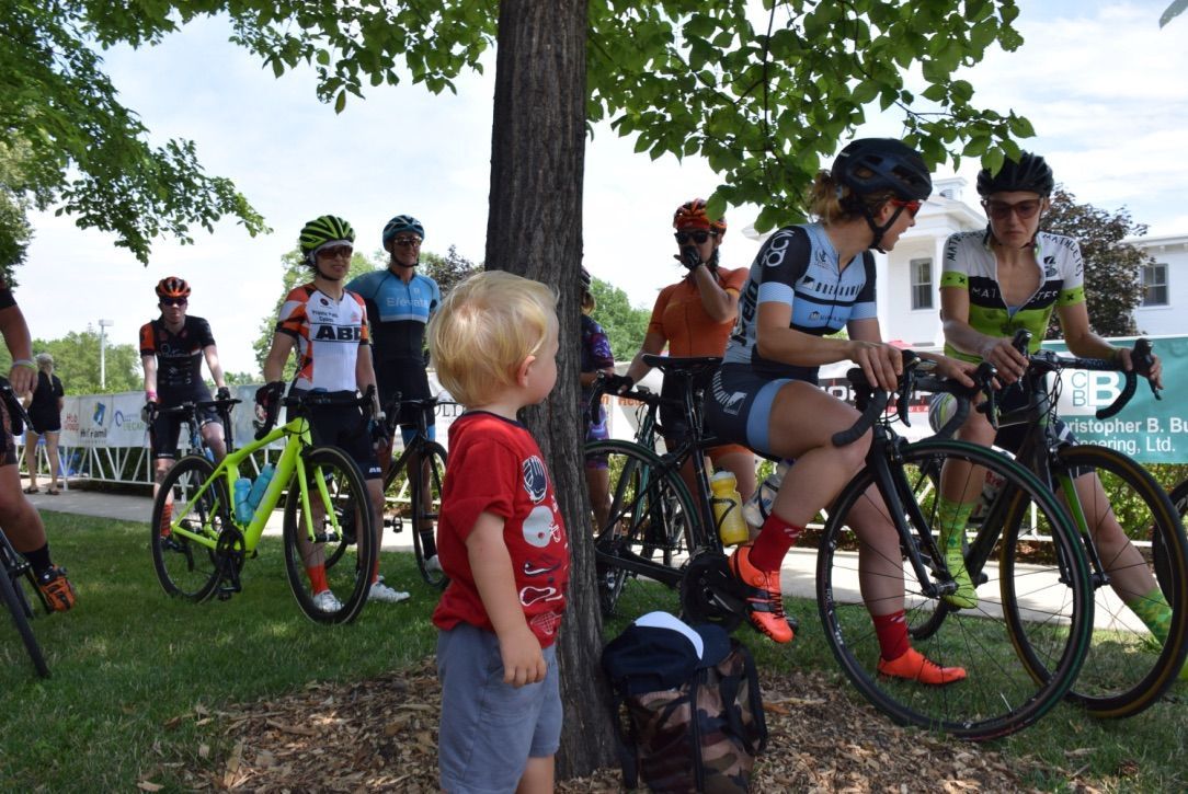 A little boy is standing under a tree next to a group of cyclists.
