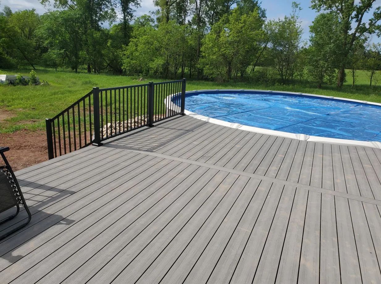 Gray deck with black railing next to an above-ground pool covered in blue. Green trees and grass in the background.