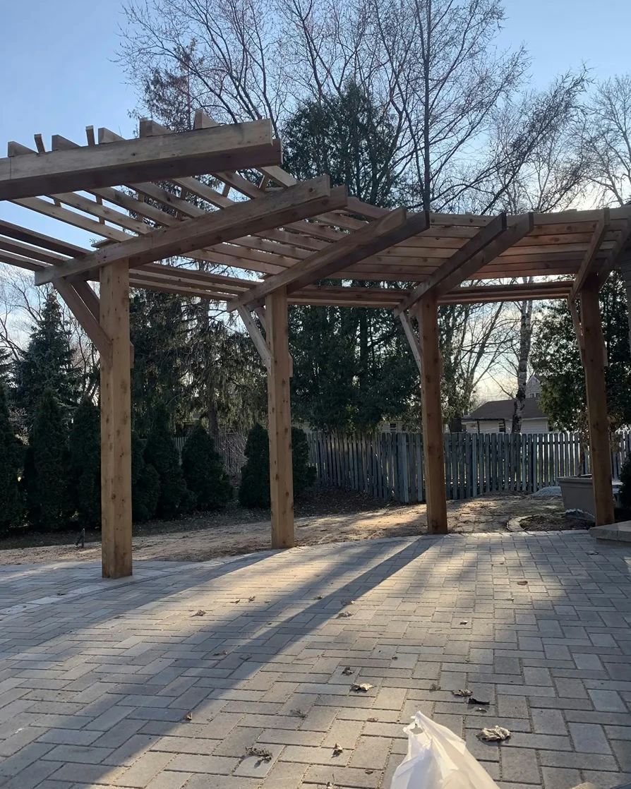 Wooden pergola over a brick patio with shadows. Trees and a fence in the background. Sunny day.