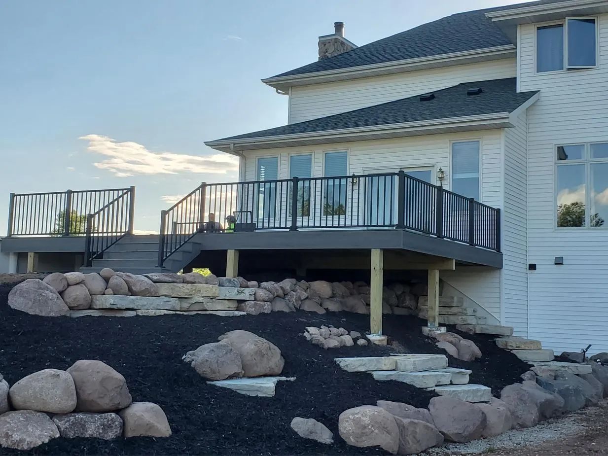 Backyard deck with black railings, connected to a white house with a dark roof, on a sloped yard with rocks.