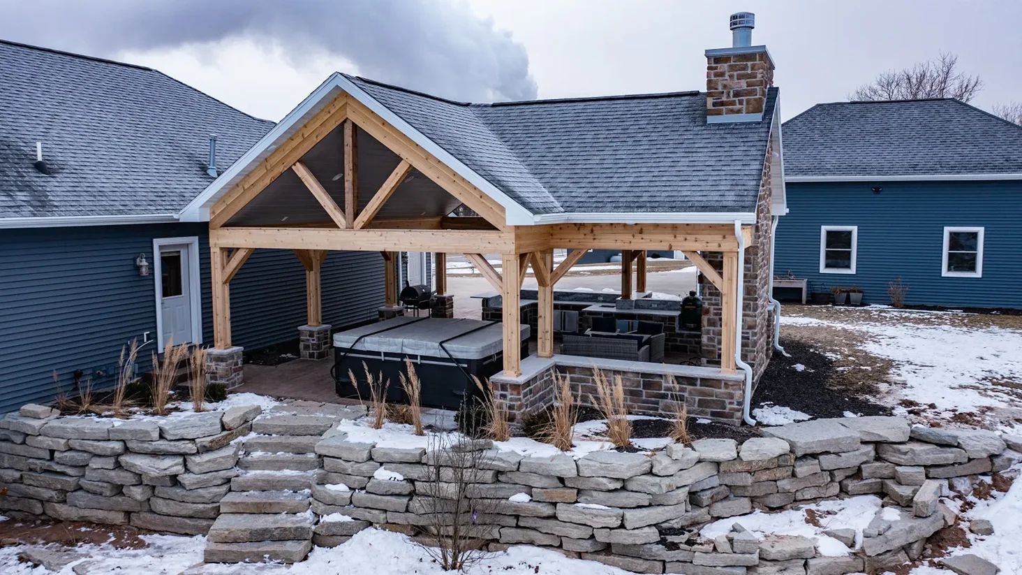 Patio with hot tub and stone accents, covered by a wooden pergola with a chimney in a snowy setting.