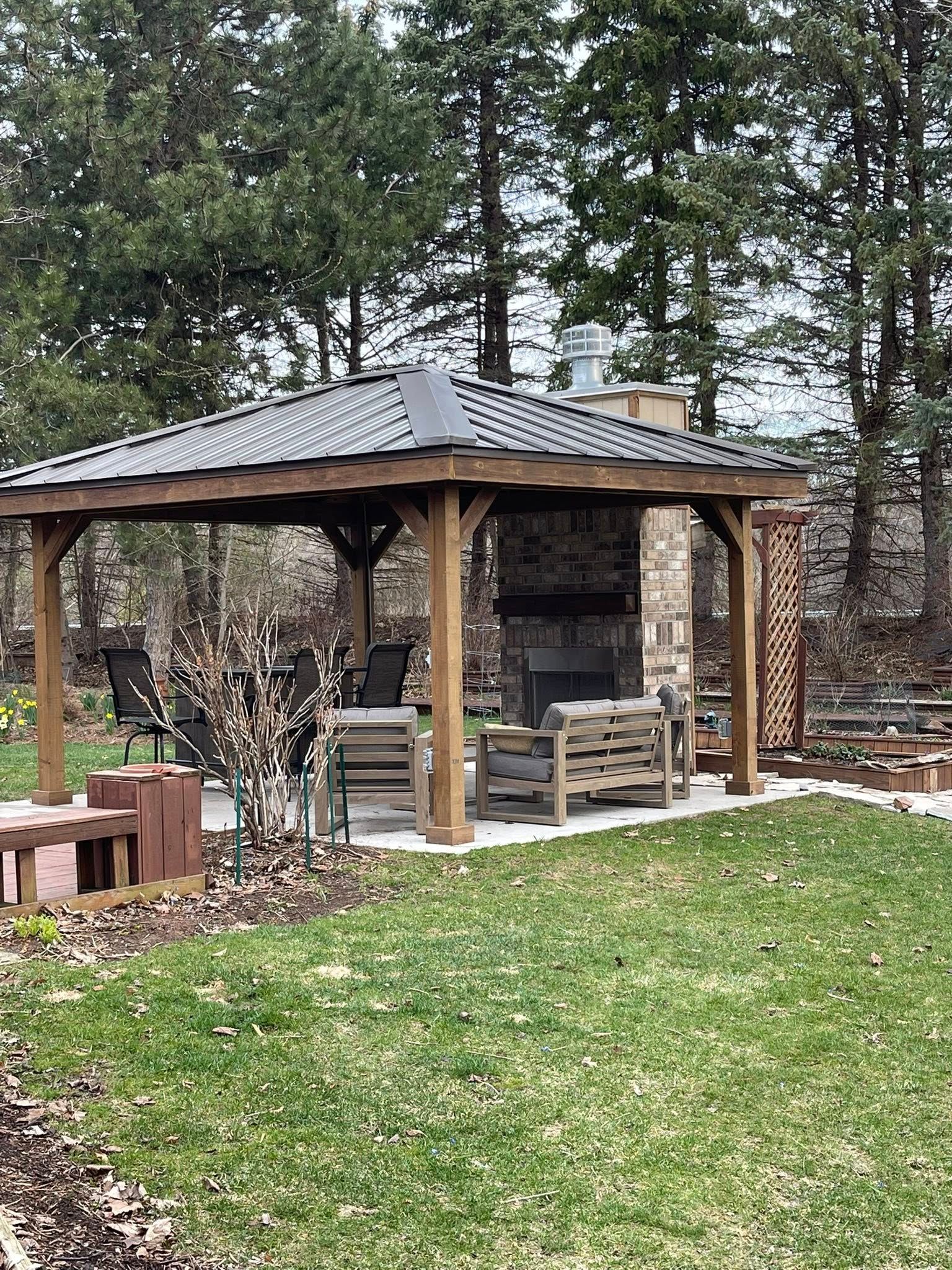 Gazebo with fireplace, wooden structure, gray roof, lawn and trees in background.