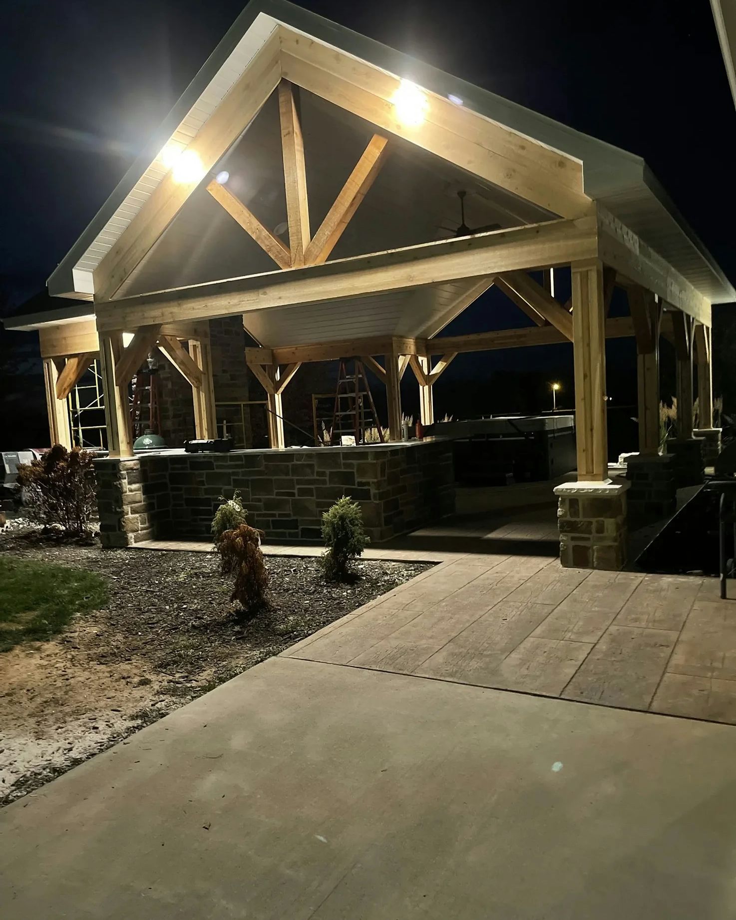 Lit outdoor wooden pavilion with stone accents and a paved patio at night.