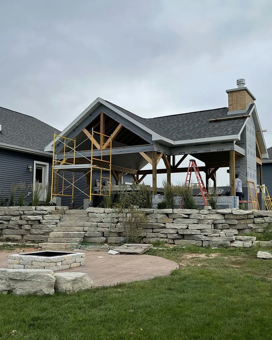 A timber frame structure under construction with a stone retaining wall and a fire pit in a yard.