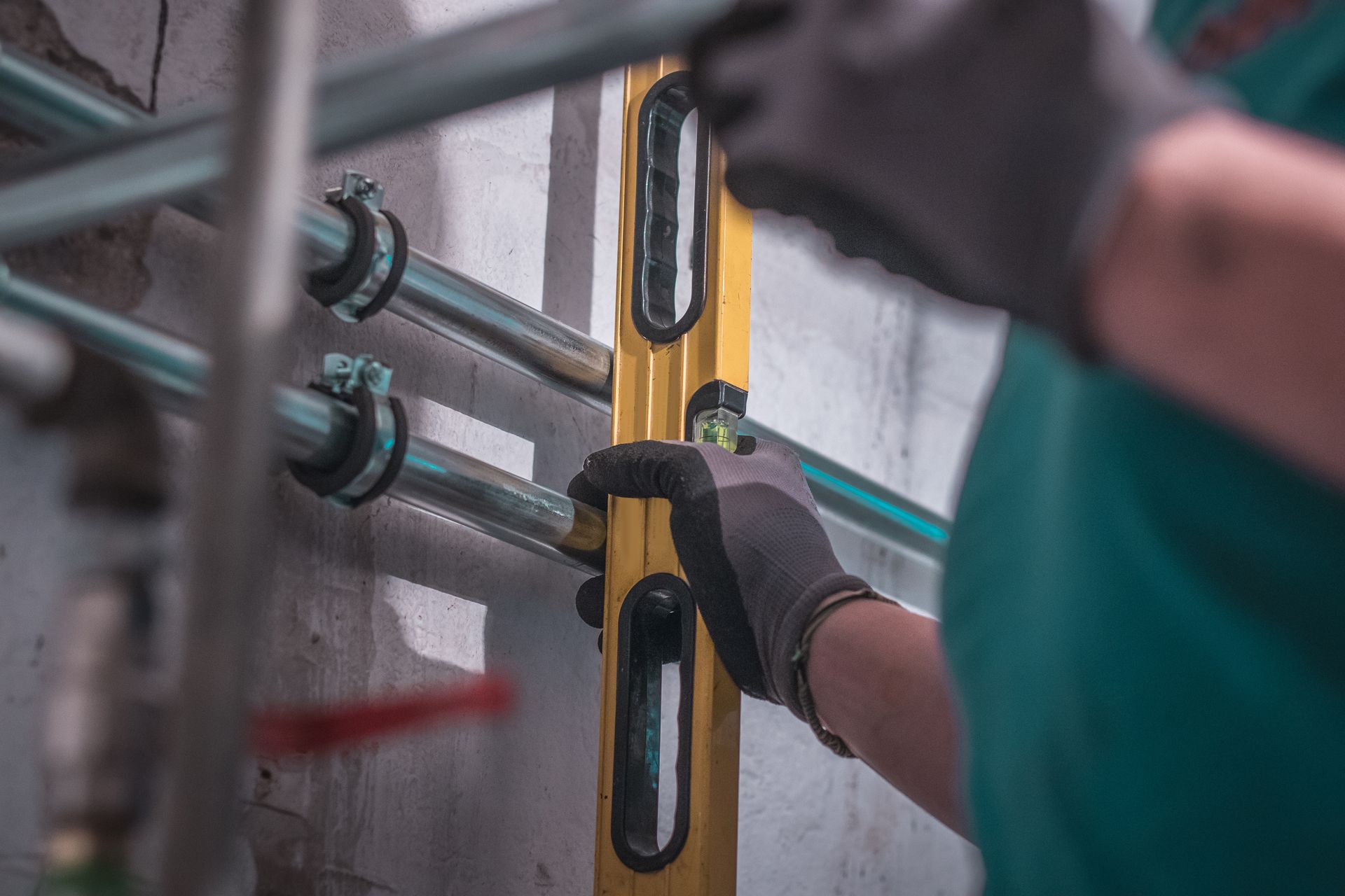 Gloved worker using a yellow spirit level to align metal pipes on a wall