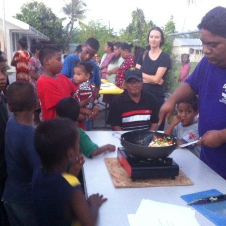 A group of people are gathered around a table with a man in a purple shirt cooking food